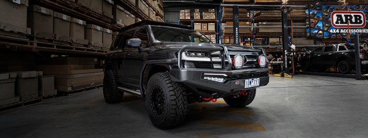 Front three-quarter view of a Toyota LandCruiser 300 Series fitted with an ARB Summit MKII bull bar, Intensity driving lights and under-vehicle protection, parked inside an ARB workshop.