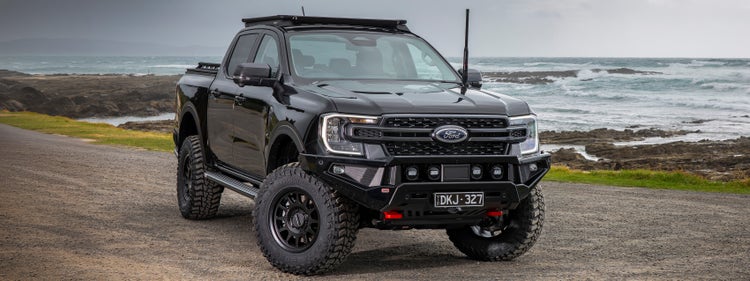 Front three-quarter view of a Ford Ranger fitted with an ARB Zenith bull bar, driving lights, roof rack and under-vehicle protection, parked beside a coastal roadway.