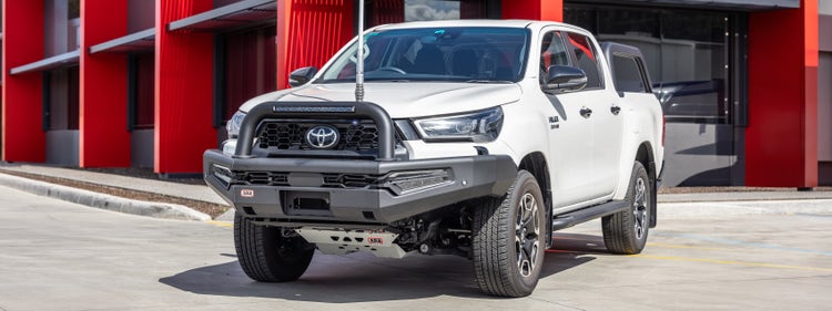 Front three-quarter view of a Toyota HiLux fitted with an ARB Summit Sahara bull bar and under-vehicle protection, parked outside an ARB store.